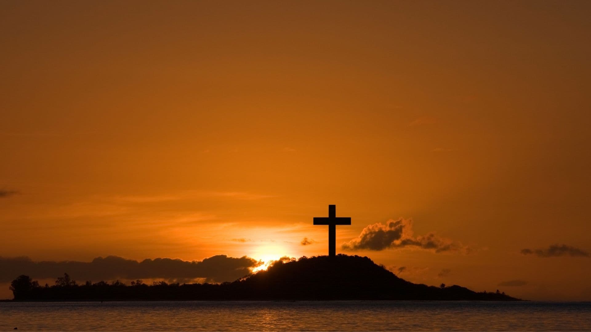 A cross silhouetted on a hill at sunset over calm water.